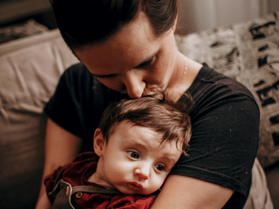 **Alt-Text:** Eine erwachsene Person hält ein Baby im Arm und küsst ihm sanft auf den Kopf, während das Kind mit nachdenklichem Blick zur Seite schaut. Die Szene wirkt ruhig und zeigt eine enge, liebevolle Bindung zwischen beiden.
