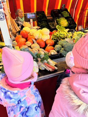 Mehrere Kinder in Winterkleidung stehen vor einem Marktstand mit Kürbis, Brokkoli und weiterem Gemüse und schauen sich die Auswahl an.