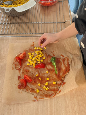 Kinderhand streut Mais auf einen ausgerollten Vollkornteig mit Tomatensoße und Gemüse.