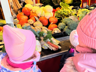 Mehrere Kinder in Winterkleidung stehen vor einem Marktstand mit Kürbis, Brokkoli und weiterem Gemüse und schauen sich die Auswahl an.