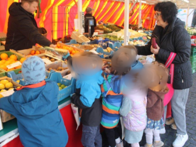Kindergruppe mit Fachkraft an einem Marktstand, gemeinsam werden Obst und Gemüse betrachtet und ausgewählt.