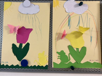 Zwei Kinderzeichnungen an einer Metallpinnwand. Die linke Zeichnung (von Alea) zeigt eine große lila Papier-Tulpe, einen rosa 3D-Schmetterling und eine kleine gelbe Maus. Die rechte Zeichnung (von Jelena) zeigt eine gelbe Tulpe, rosa Schmetterlinge und eine grüne Maus, dazu farbige Regenbogen- und Wolkenzeichnungen.