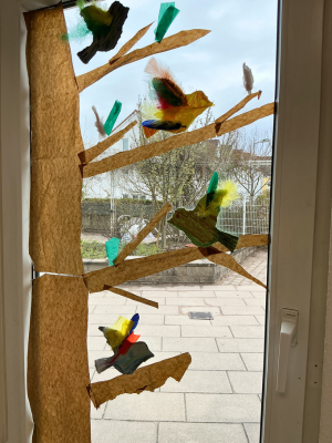 Ein Fenster mit Blick auf einen Hof. Am Glas sind ein brauner Papier-Baumstamm und Äste befestigt. Auf den Ästen sitzen mehrere kleine Papier-Vögel, die mit bunten Federn (grün, gelb, rot, blau) dekoriert sind.