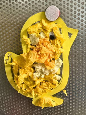 Eine Nahaufnahme einer Kinderbastelarbeit: Ein ausgeschnittenes gelbes Papier-Küken, dessen Körper mit kleinen Knubbeln aus gelbem und weißem Krepppapier beklebt ist. Es hat ein Wackelauge.