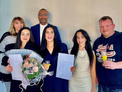 Sechs Personen mit Urkunden und Blumen stehen nebeneinander und lächeln in die Kamera, vermutlich bei einer Ehrung oder einem Abschluss.