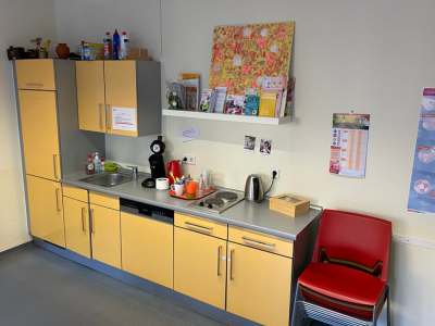 Kleine Küchenzeile im Elterncafé der AWO Karlsruhe mit Spüle, Kaffeemaschine und roten Stapelstühlen. Die bisherige Küche bot nur eingeschränkte Möglichkeiten für gemeinsames Kochen oder Backen.