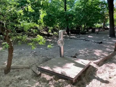 Neu hergerichteter Sandbereich im Außengelände der Kita am Weinbrennerplatz. Eine fest installierte Wasserpumpe aus Metall steht auf einer Holzplattform im frisch geglätteten Sand, umgeben von Bäumen.