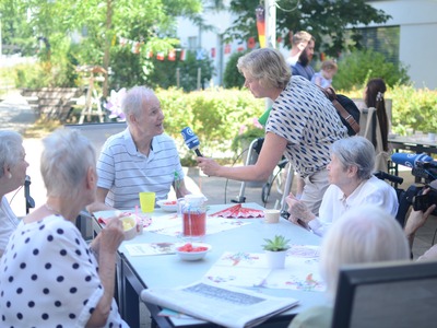 Eine Reporterin interviewt einen Bewohner im Garten eines Seniorenzentrums. Weitere Seniorinnen und Senioren sitzen an einem Tisch und genießen kühle Getränke.