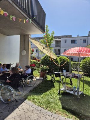 Bewohner*innen des Seniorenzentrums Knielingen sitzen auf der Terrasse unter Sonnenschirmen beim Erdbeerfest; im Hintergrund bunte Girlanden und ein Servierwagen.