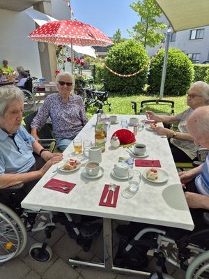 Vier Bewohner*innen sitzen an einem Tisch im Garten des Seniorenzentrums Knielingen; im Hintergrund roter Tupfen-Sonnenschirm und bunte Girlanden.
