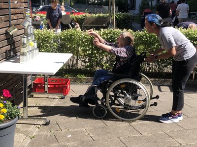 Bewohnerin im Rollstuhl zielt mit einem Tennisball auf gestapelte Dosen beim Dosenwurf-Spiel; eine Mitarbeiterin unterstützt sie beim Sportfest im AWO Anna Leimbach Haus Karlsruhe.
