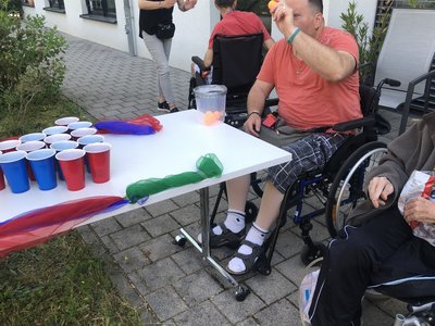 .Bewohner im Rollstuhl wirft einen Ball in einen Behälter an einer Spielstation; auf dem Tisch bunte Becher und Tücher beim Sportfest im AWO Anna Leimbach Haus Karlsruhe.