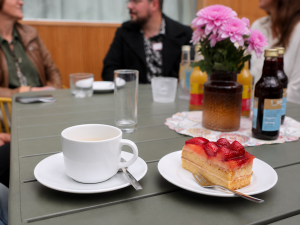 Eine weiße Tasse steht zusammen mit einem Stück Erdbeerkuchen auf einem Tisch mit Blumen. Im Hintergrund unterhalten sich Menschen.