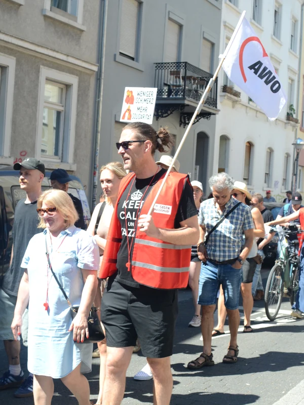 Teilnehmende einer Demonstration auf einer Straße; im Vordergrund trägt ein Mann in roter Warnweste eine weiße AWO-Fahne, daneben und dahinter gehen weitere Demonstrierende mit.