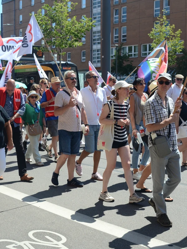 Gruppe von Demonstrierenden auf einer breiten Straße mit mehreren AWO-Fahnen und einer Regenbogenflagge; die Teilnehmenden gehen gemeinsam bei hellem Sommerwetter.