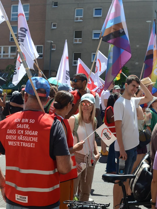 Demonstrierende stehen auf einer Straße zwischen AWO-Fahnen und bunten Pride-Flaggen; im Vordergrund sind Personen in roten Warnwesten und eine kleine AWO-Handfahne zu sehen.