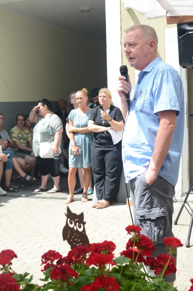 Ein Mann steht mit einem Mikrophon vor roten Blumen. Dahinter stehen Menschen und hören ihm zu.