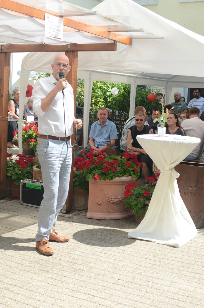 Ein Mann steht links mit einem Mikrophon. Daneben ein Stehtisch und Menschen auf Bierbänken unter einem Pavillon.