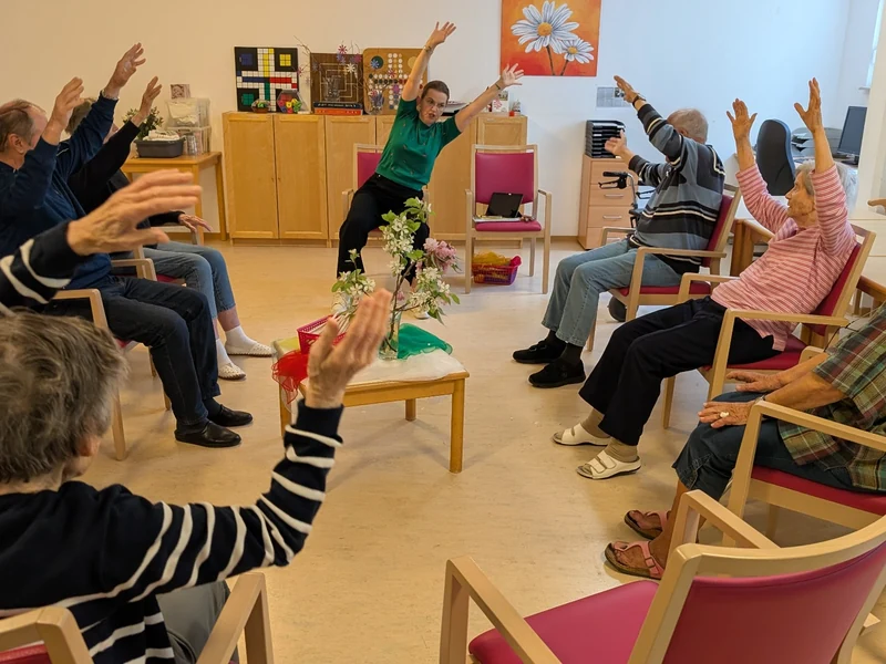 Senior*innen sitzen im Stuhlkreis und heben während einer angeleiteten Bewegungseinheit gemeinsam die Arme.