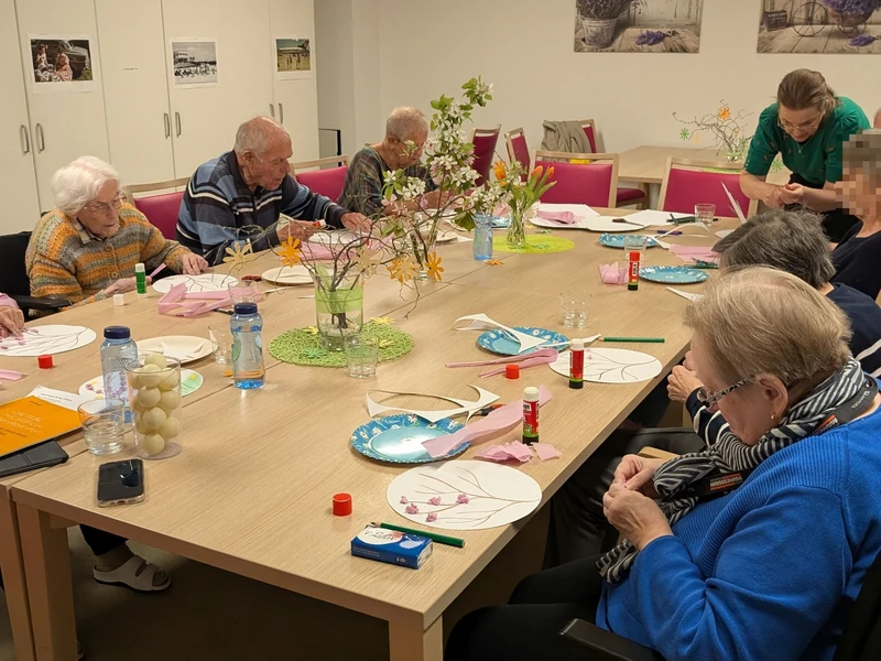 Senior*innen gestalten gemeinsam an einem großen Tisch Papierbilder mit Blüten und Zweigen.