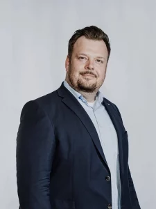 Porträt eines Mannes im dunklen Sakko und hellblauen Hemd vor einem neutralen Hintergrund.