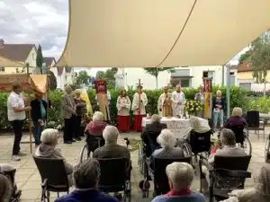 Geistliche Begleitende, Fahnen und Bewohner*innen bei der Fronleichnamsfeier auf der Terrasse.