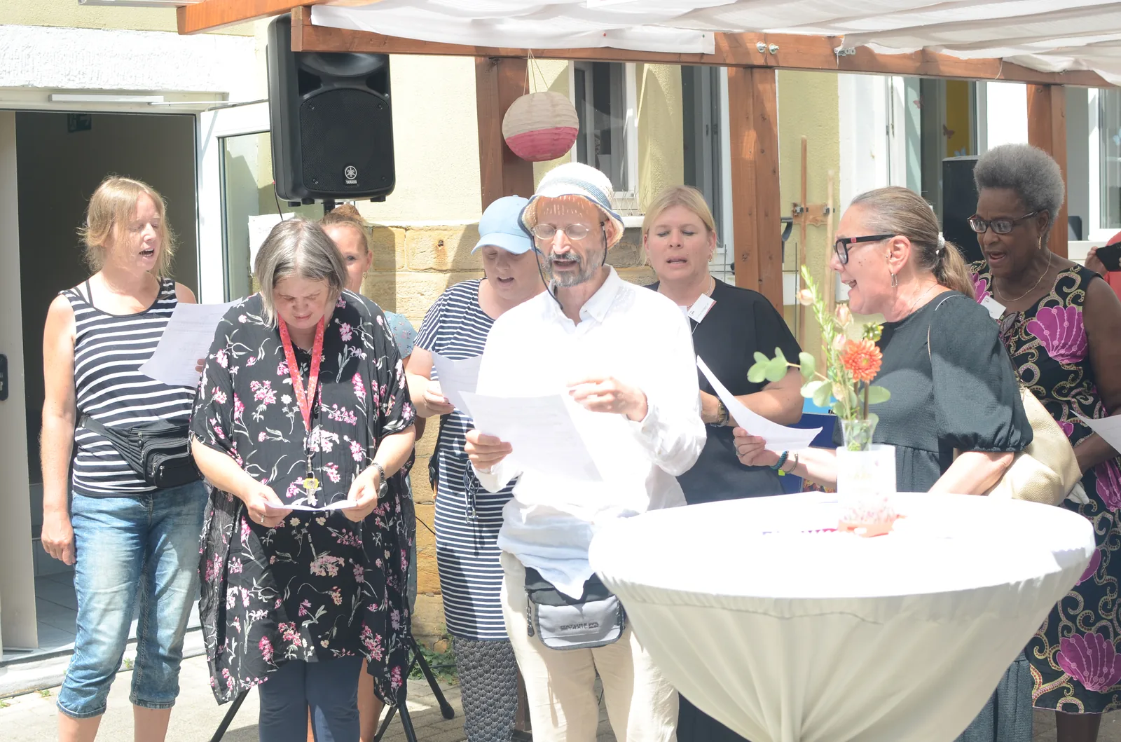 Menschen stehen im Sonnenschein an einem Stehtisch und singen.