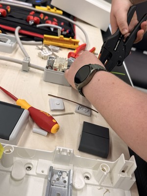 Eine Person isoliert mit einer Zange ein Kabel ab; auf dem Arbeitstisch liegen Werkzeuge und Bauteile.