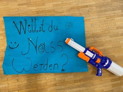 Eine Wasserpistole liegt neben einem blauen Plakat mit der Aufschrift „Willst du nass werden?“.