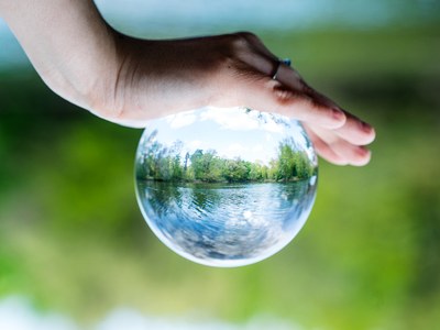 Eine Hand hält eine Glaskugel, in der sich See und Bäume spiegeln.