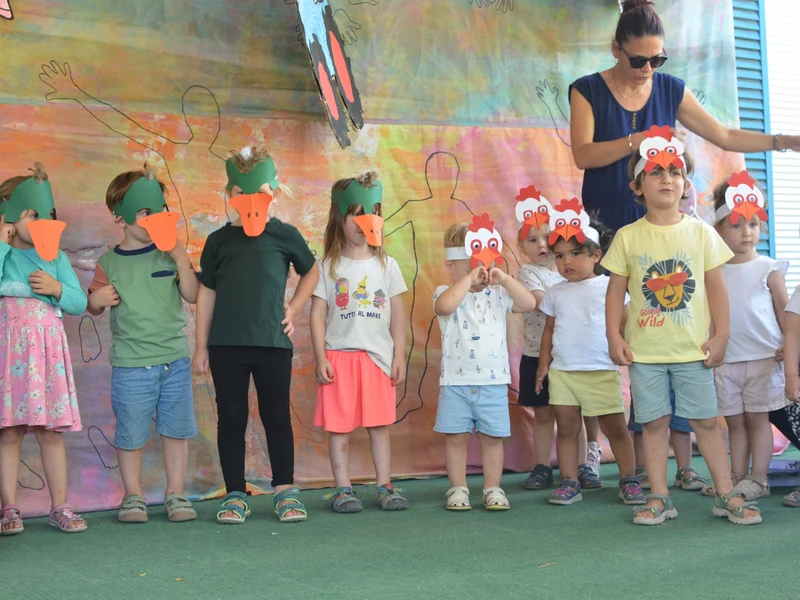 Kinder mit selbst gebastelten Enten- und Hühnermasken stehen bei der Sommerfest-Aufführung vor einer bunt bemalten Kulisse.