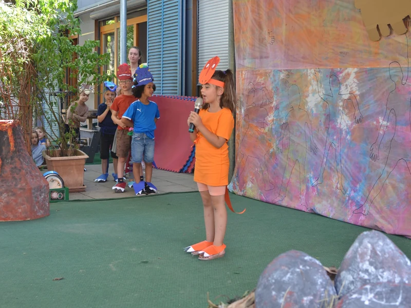 Ein Kind spricht beim Sommerfest mit einem Mikrofon, während weitere Kinder mit Dinosauriermasken im Hintergrund warten.