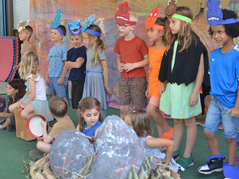 Kinder mit selbst gebastelten Dinosauriermasken stehen während der Aufführung vor einer bunt bemalten Kulisse.