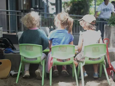 Drei Kinder sitzen mit dem Rücken zur Kamera auf kleinen grünen Stühlen im Außengelände einer Kita.