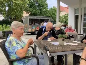 Eine Bewohnerin und ein Bewohner sitzen an einem Terrassentisch und essen Eis, im Hintergrund ist der Eiswagen zu sehen.