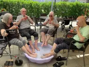 Mehrere Bewohnerinnen in Rollstühlen sitzen im Kreis um eine kleine Wasserschale, in der sie die Füße kühlen, eine Frau isst Wassermelone.