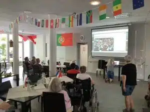 Bewohner*innen sitzen im Gemeinschaftsraum vor einer Leinwand mit WM-Bildern, darüber hängen Fähnchen verschiedener Länder von der Decke.