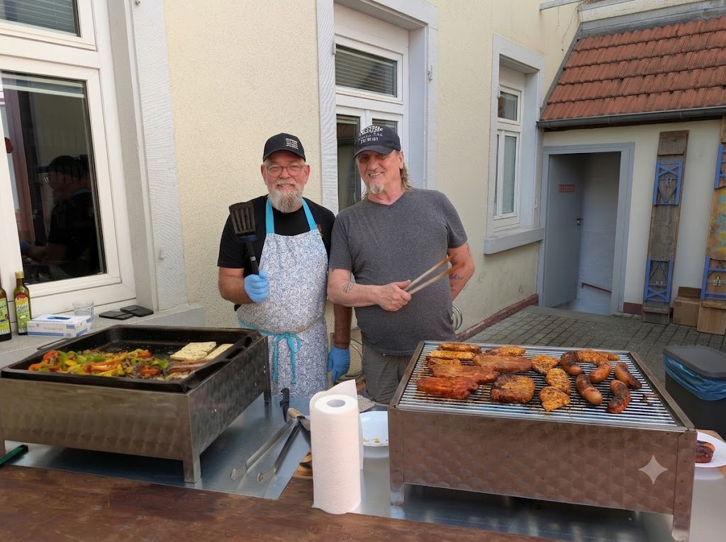 Zwei Männer stehen vor zwei Grilgeräten auf denen Fleischstücke liegen.