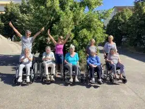 Acht Frauen posieren für ein Gruppenfoto unter einem Baum, fünf davon im Rollstroller, zwei Betreuerinnen strecken lachend die Arme in die Höhe.