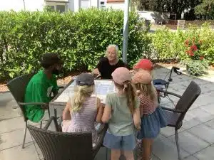 Fünf Kinder mit Sonnenhüten sitzen an einem Gartentisch einer älteren Frau gegenüber, im Hintergrund ein junger Mann mit Cap, umgeben von Hecken und Blumen.