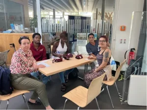 Besucher*innen und Mitarbeitende sitzen mit Getränken und Blutdruckmessgeräten an einem Tisch.