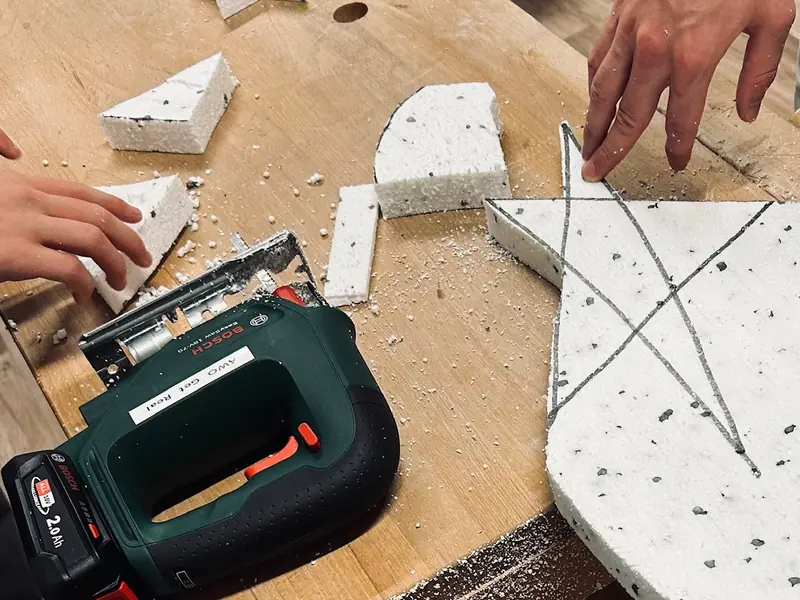Hände führen eine Stichsäge entlang angezeichneter Formen auf einer Styroporplatte.