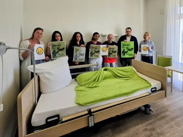 Eine Gruppe steht hinter einem leeren Pflegebett mit grüner Decke; mehrere Personen halten Aktionsplakate.