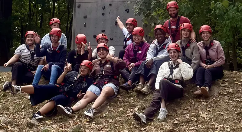 Das Team der Kita Mikado sitzt in Kletterausrüstung vor einer Kletterwand im Wald.