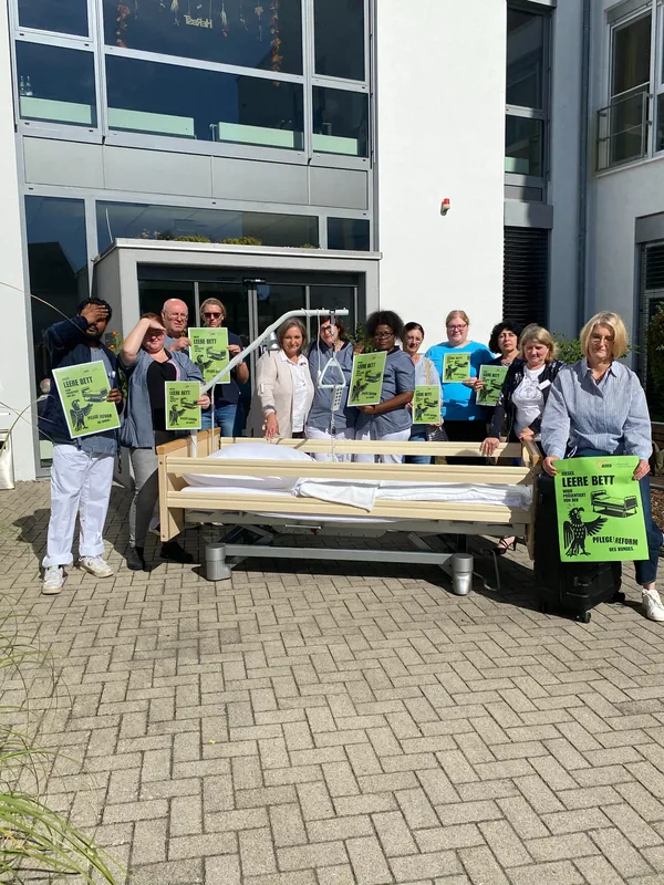 Mitarbeitende stehen vor einem Gebäude hinter einem leeren Pflegebett und halten grüne Plakate zur Aktion „Das leere Pflegebett“.