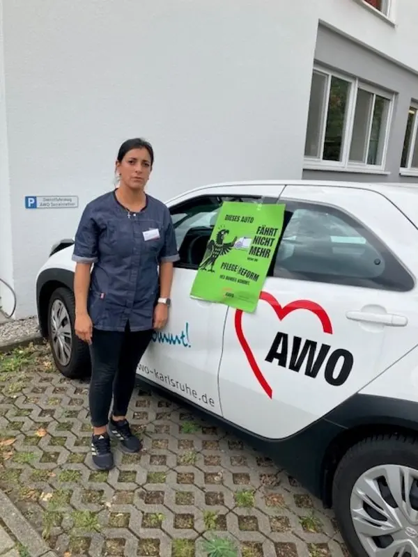 Eine Mitarbeiterin der ambulanten Pflege steht neben einem AWO-Dienstwagen; an der Fahrzeugseite ist ein grünes Aktionsplakat angebracht.