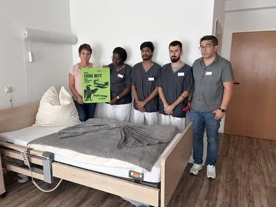 Fünf Mitarbeitende stehen neben einem leeren Pflegebett; eine Person hält ein grünes Aktionsplakat.