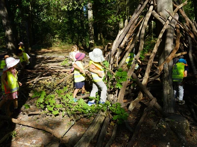 Kinder mit gelben Warnwesten stehen an einer aus Ästen errichteten Waldhütte; zwei Kinder tragen einen Ast.