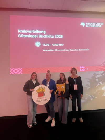 Vier Frauen stehen vor einer Bühnenprojektion zur Preisverleihung des Gütesiegels Buchkita 2026.