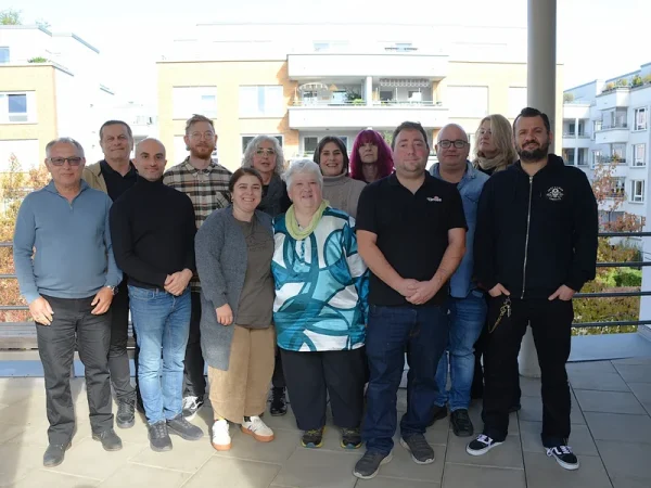 Zwölf Personen stehen in zwei Reihen für ein Gruppenfoto auf einer Terrasse. Im Hintergrund sind Wohngebäude zu sehen.